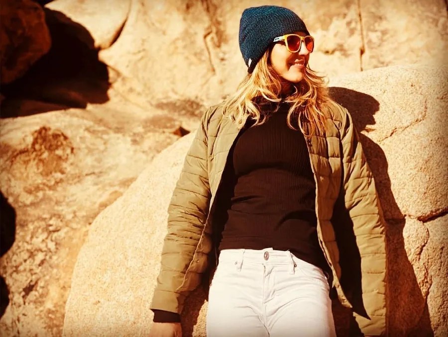 Kat in a beanie and sunglasses leaning against desert rocks